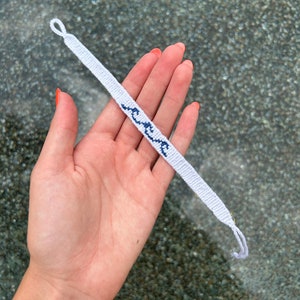 May include: White friendship bracelet with blue woven waves. The bracelet is held in a hand.