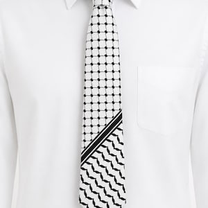 May include: A necktie featuring a black and white keffiyeh pattern, displayed against a crisp white collared dress shirt. The tie showcases a grid-like dot design on the upper portion and a traditional wave-like pattern on the lower section.