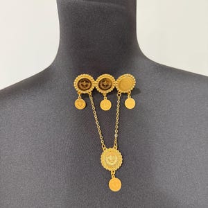 May include: A gold-colored necklace and brooch set. The brooch features three circular elements with dangling coin-shaped charms. A chain connects the brooch to a larger circular pendant with a dangling coin.