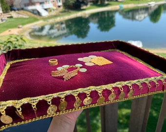 May include: A rectangular tray covered in deep burgundy velvet fabric with gold metallic trim and dangling gold coin accents. The tray holds a gold ring, a gold leaf-shaped bracelet, and several small gold coins scattered across the surface.