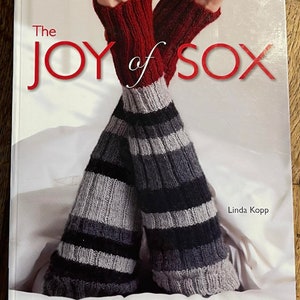 May include: Book cover with the title "The Joy of Sox" in red lettering. The cover features a pair of striped socks in grey and black with red cuffs. The author's name, Linda Kopp, is printed in black at the bottom right corner. The book title also includes the words "30+ Must-Knit Designs".