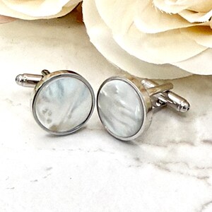 May include: A pair of round cufflinks featuring a polished silver-tone metal frame and a smooth, iridescent white mother-of-pearl inlay. Each cufflink has a standard hinged toggle mechanism on the back for securing through shirt buttonholes.