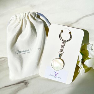 May include: A silver-toned keychain featuring a circular pendant with a white iridescent mother-of-pearl inlay, attached to a white display card with the text Handmade Anniversary and a small heart logo, next to a white fabric drawstring pouch.