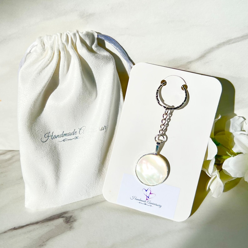 May include: A silver-toned keychain featuring a circular pendant with a white iridescent mother-of-pearl inlay, attached to a white display card with the text Handmade Anniversary and a small heart logo, next to a white fabric drawstring pouch.