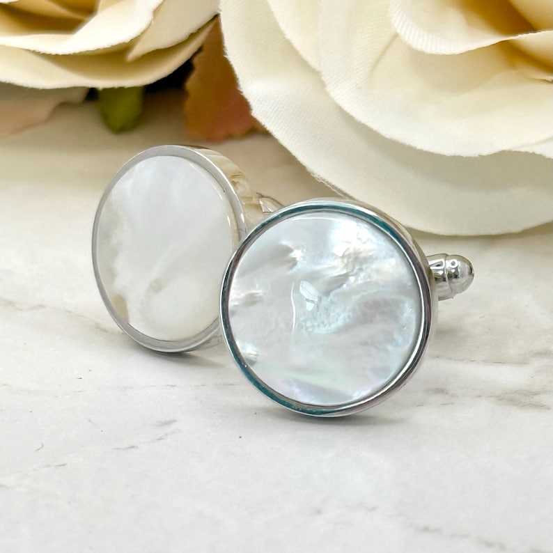 May include: A pair of round cufflinks featuring a polished silver-tone metal frame and a circular inlay of iridescent white mother-of-pearl. The surface of the inlay displays a natural, swirling texture with subtle pearlescent highlights.