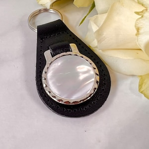 Puede incluir: Un llavero de cuero negro con una incrustaci&oacute;n circular de n&aacute;car blanco iridiscente engastada en un marco de metal plateado con bordes festoneados. La base de cuero tiene forma de l&aacute;grima con costuras visibles y una anilla plateada.