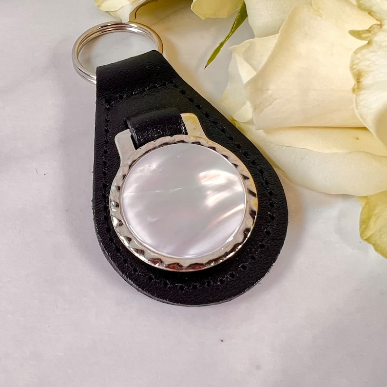 Puede incluir: Un llavero de cuero negro con una incrustaci&oacute;n circular de n&aacute;car blanco iridiscente engastada en un marco de metal plateado con bordes festoneados. La base de cuero tiene forma de l&aacute;grima con costuras visibles y una anilla plateada.
