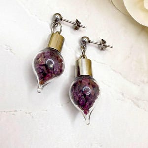 May include: Earrings with glass vials filled with deep red gemstones. The teardrop-shaped vials have silver-coloured metal caps and posts. The gemstones are a rich, dark red colour, creating a striking contrast with the metal.