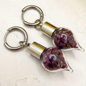 May include: A pair of earrings featuring small glass vials filled with deep red gemstones. The vials have silver-colored metal caps and are attached to silver hoop earrings. The gemstones appear to be ruby.