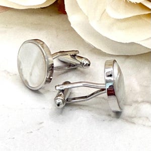 May include: A pair of silver-toned cufflinks featuring circular faces with a smooth, iridescent white mother-of-pearl inlay. Each cufflink has a polished metal frame and a standard hinged toggle mechanism for securing through shirt buttonholes.