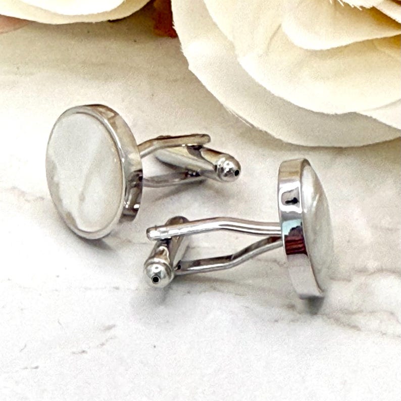 May include: A pair of silver-toned cufflinks featuring circular faces with a smooth, iridescent white mother-of-pearl inlay. Each cufflink has a polished metal frame and a standard hinged toggle mechanism for securing through shirt buttonholes.