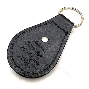 May include: A teardrop-shaped black leather keychain featuring a silver metal split ring and a silver rivet. The surface is embossed with the text: Adrian, With love, 25 August 2001. The edges are finished with black stitching around the perimeter.