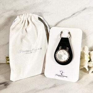 May include: A black leather keychain featuring a circular mother-of-pearl inlay set in a silver-toned metal frame, attached to a silver ring. The keychain is mounted on a white display card with the text Handmade Anniversary and a heart logo.