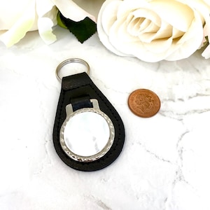 Puede incluir: Un llavero de cuero negro con forma de l&aacute;grima que presenta un marco circular de metal plateado con una incrustaci&oacute;n de n&aacute;car blanco. Un anillo de metal plateado est&aacute; unido a la parte superior. Una moneda brit&aacute;nica de cobre est&aacute; al lado.