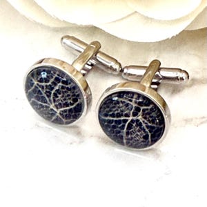 May include: A pair of silver cufflinks with a dark blue and white patterned design. The cufflinks have a round face with a detailed, web-like pattern. The metal has a shiny, polished finish.