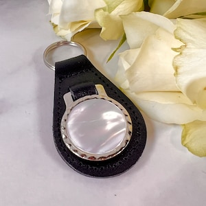 Puede incluir: Un llavero de cuero negro con una incrustaci&oacute;n circular de n&aacute;car blanco iridiscente engastada en un marco de metal plateado con bordes festoneados. La pieza de cuero en forma de l&aacute;grima est&aacute; unida a una anilla circular de metal plateado.