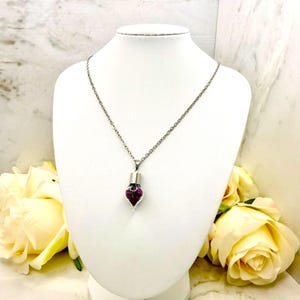May include: A silver necklace featuring a teardrop-shaped pendant with a dark purple gem. The necklace is displayed on a white mannequin. Yellow roses are visible in the background, adding a touch of colour.