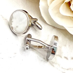 May include: A pair of round cufflinks featuring a polished silver-tone metal frame and a smooth, iridescent white mother-of-pearl inlay. The cufflinks have a standard hinged toggle mechanism on the back, designed to secure through shirt cuffs.