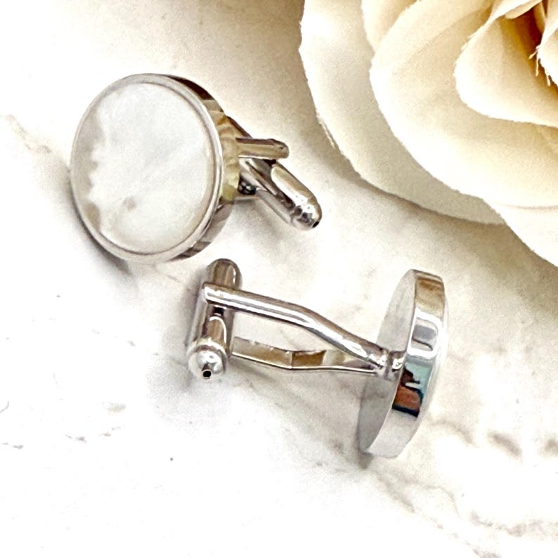 May include: A pair of round cufflinks featuring a polished silver-tone metal frame and a smooth, iridescent white mother-of-pearl inlay. The cufflinks have a standard hinged toggle mechanism on the back, designed to secure through shirt cuffs.