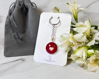 May include: A red and white heart keychain with a silver chain and ring. The keychain is on a white card with the text "Handmade Anniversary" printed on it. The keychain is next to a gray velvet bag and white flowers.
