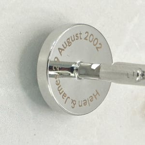 May include: A circular silver-toned cufflink featuring a polished metallic finish. The face of the cufflink is engraved with the text "Helen & James 29 August 2002" in a clean, sans-serif font, centered around the central post attachment.