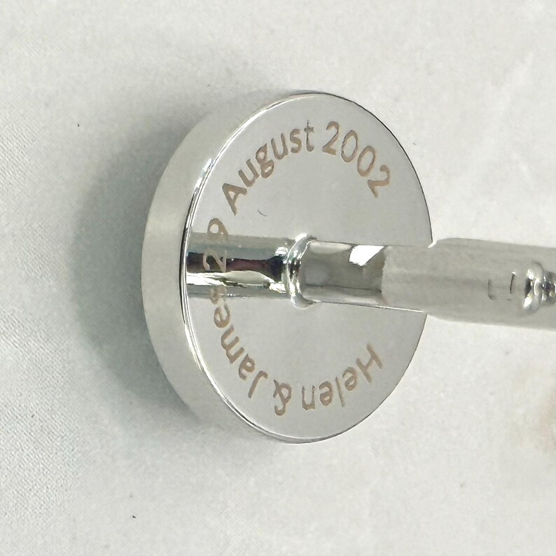 May include: A circular silver-toned cufflink featuring a polished metallic finish. The face of the cufflink is engraved with the text "Helen & James 29 August 2002" in a clean, sans-serif font, centered around the central post attachment.