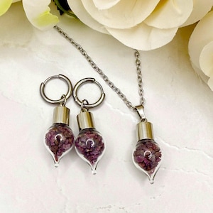 May include: A jewelry set featuring a necklace and hoop earrings. Each piece has a clear glass vial filled with small, deep pink gemstones. The vials are capped with silver-toned metal and suspended from silver chains and hoops.