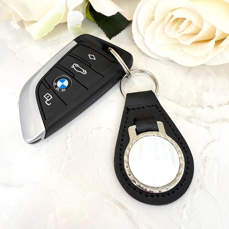 Puede incluir: Un mando de coche BMW negro con detalles plateados y un logotipo circular, unido a un llavero de cuero negro con forma de l&aacute;grima. El llavero presenta un borde de metal plateado que rodea un centro circular blanco en blanco.