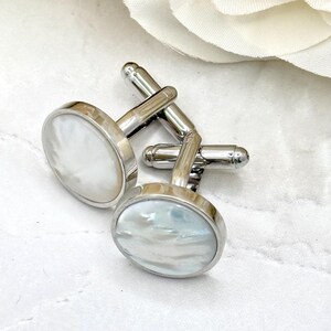 May include: A pair of round cufflinks featuring polished silver-tone metal frames and iridescent white mother-of-pearl inlays. Each cufflink has a standard swivel toggle mechanism on the back, showcasing a smooth, reflective surface finish.