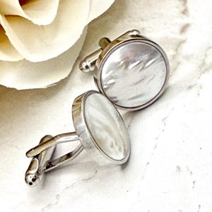 May include: A pair of round cufflinks featuring a polished silver-tone metal frame and a smooth, iridescent white mother-of-pearl inlay. Each cufflink has a circular face with a reflective surface and a standard hinged toggle mechanism on the back.