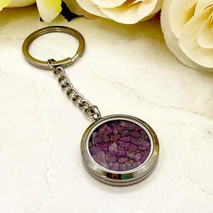May include: A silver keyring with a circular pendant containing small, dark pink gemstones. The pendant is connected to a chain and a ring. The background shows a marble surface and a blurred floral element.