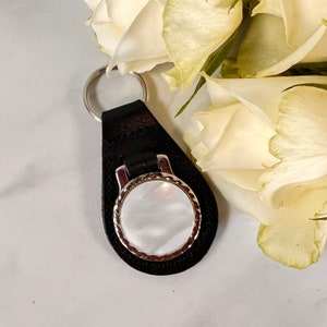 Puede incluir: Un llavero de cuero negro con un marco circular de metal plateado y una incrustaci&oacute;n de n&aacute;car blanco iridiscente. El componente circular est&aacute; unido a una base de cuero negro en forma de l&aacute;grima con una anilla de metal plateado.