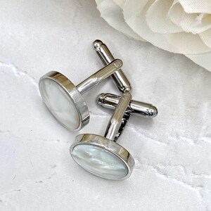May include: A pair of round cufflinks featuring a polished silver-tone metal frame and a smooth, iridescent white mother-of-pearl inlay. Each cufflink has a sturdy T-bar swivel mechanism attached to the back for secure fastening on a shirt cuff.