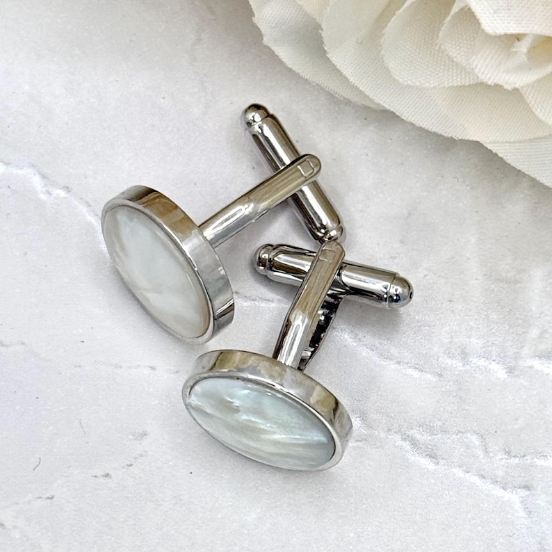 May include: A pair of round cufflinks featuring a polished silver-tone metal frame and a smooth, iridescent white mother-of-pearl inlay. Each cufflink has a sturdy T-bar swivel mechanism attached to the back for secure fastening on a shirt cuff.
