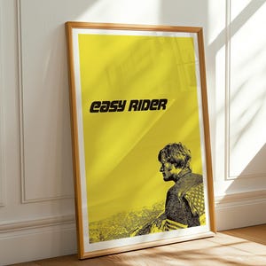 Könnte beinhalten: Ein gerahmtes Poster mit gelbem Hintergrund und den schwarzen Worten "easy rider". Das Poster zeigt ein Schwarz-Weiß-Bild einer Person im Profil. Der Rahmen ist aus Holz.