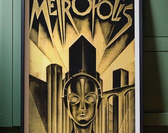 May include: A framed art print of the movie "Metropolis". The poster features a stylized robot figure in the foreground with a cityscape and the film's title in gold lettering against a black and gold background.