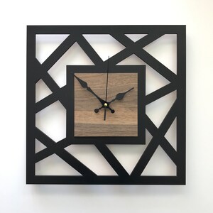 Peut inclure: Une horloge murale carrée noire avec un design géométrique. Le cadran de l'horloge est un carré en bois avec des aiguilles noires et un centre doré.