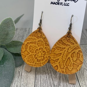 May include: A pair of mustard yellow leather earrings with a lace pattern. The earrings are teardrop shaped and have gold-toned hooks.