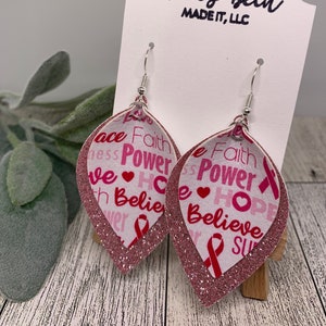 May include: Pink glitter dangle earrings with a white background featuring the words "Faith", "Power", "Hope", and "Believe" with pink ribbon accents. The earrings are displayed on a white card with the text "Molly Beth Made It, LLC".