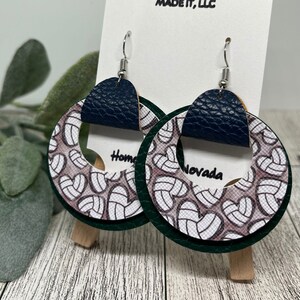 May include: A pair of green, white, and navy blue volleyball-themed earrings. The earrings are made of leather and have a circle shape with a smaller circle cut out in the center. The earrings are labeled "Home" and "Nevada".