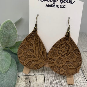 Könnte beinhalten: Ein Paar braune Lederohrringe mit einem Spitzenmuster. Die Ohrringe hängen an einem Holzständer mit dem Text "Molly Beth Made It, LLC" auf der Vorderseite.