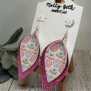 May include: Dangle earrings with a layered design. The inner layer has a pattern of colourful donuts on a white background. The outer layer is a bright pink glitter. The earrings are displayed on a white card with the text "Molly Beth MADE IT, LLC".
