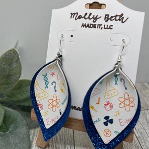 May include: A pair of blue glitter dangle earrings with a white leather teardrop shape. The teardrop is printed with science-themed illustrations, including a beaker, atom, DNA strand, and the equation E=MC2.