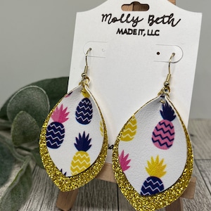 Puede incluir: Pendientes de aro de oro con purpurina con fondo blanco y estampado de piña. Los pendientes están etiquetados con "Molly Beth Made It, LLC".