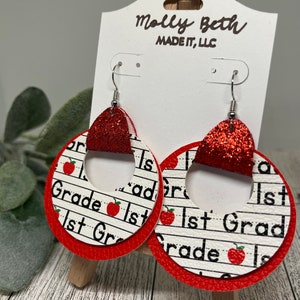 May include: Red and white dangle earrings with a red glitter accent. The earrings feature a layered design with a white circle with the text "1st Grade" repeated and a red circle around the white circle.