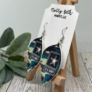 May include: A pair of green, black and white camouflage leaf-shaped earrings with a silver hook and a white star on a black U.S. Army patch.