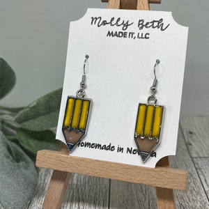 May include: A pair of pencil-shaped earrings with yellow and brown enamel, outlined in silver. The earrings are displayed on a white card with the text "Molly Beth MADE IT, LLC" and "Homemade in Nova".