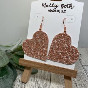 May include: Rose gold glitter heart shaped dangle earrings with a copper wire hook. The earrings are displayed on a white card with the text "Molly Beth Made It, LLC".