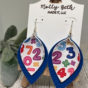Puede incluir: Un par de pendientes colgantes azules y blancos con un patrón de números coloridos. Los pendientes están hechos de cuero y tienen forma de lágrima. Los pendientes están en una tarjeta blanca con el texto "Molly Beth Made It, LLC".