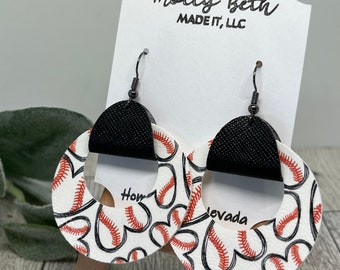 Puede incluir: Pendientes colgantes con diseño circular blanco y patrón de corazones con costuras de béisbol en negro y naranja. Cuentan con una pieza superior semicircular negra texturizada y ganchos de metal oscuro, presentados en una tarjeta blanca.
